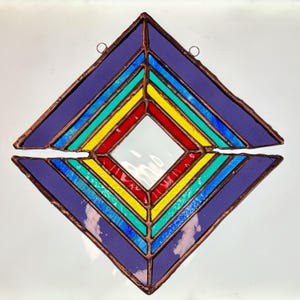 May include: A stained glass suncatcher in a diamond shape, featuring a vibrant array of colors. The design includes purple, blue, turquoise, yellow, and red glass panels, with a copper-colored frame. Two small rings are attached for hanging.