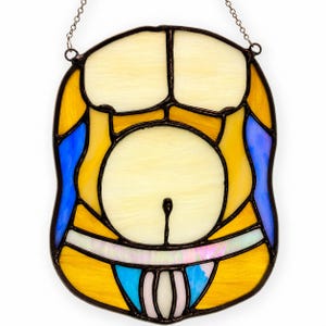 May include: A stained glass suncatcher shaped like a torso, featuring cream-colored glass for the chest and abdomen, accented with yellow and blue glass panels. The piece is outlined in dark metal solder and includes a metal chain for hanging.