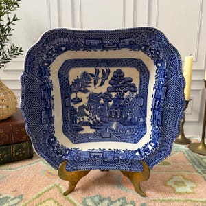 May include: A square, blue and white ceramic serving dish with a scalloped edge. The center features a detailed landscape scene. The dish is displayed on a gold-colored metal stand, showcasing a classic Willow pattern.