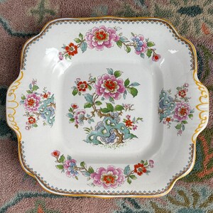 Pode incluir: Prato quadrado de cer&acirc;mica branca com um desenho floral com flores rosa, vermelhas e azuis. O prato tem uma borda ondulada com acabamento dourado e al&ccedil;as. O centro do prato tem um arranjo floral detalhado.