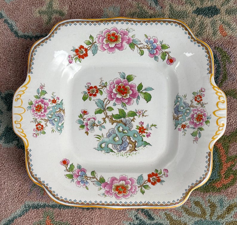 Pode incluir: Prato quadrado de cer&acirc;mica branca com um desenho floral com flores rosa, vermelhas e azuis. O prato tem uma borda ondulada com acabamento dourado e al&ccedil;as. O centro do prato tem um arranjo floral detalhado.