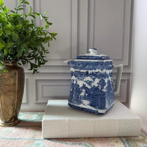 May include: A square-shaped ceramic teapot featuring a blue and white willow pattern with a lid and a side handle. The teapot displays detailed illustrations of bridges, pagodas, and trees, and it is positioned on a thick, light-colored rectangular book.