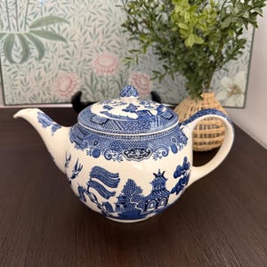 May include: A white and blue ceramic teapot with a lid, featuring a detailed blue willow pattern. The teapot has a curved spout and handle, and a small knob on the lid. The design includes trees, buildings, and birds.