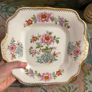 Pode incluir: Prato quadrado de cer&acirc;mica branca com um desenho floral. O prato apresenta flores rosa, vermelhas e azuis com folhas verdes e uma borda amarela ondulada. O prato tem uma borda decorativa.