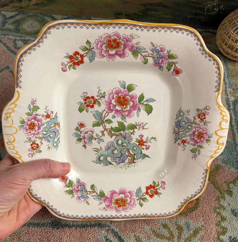 Pode incluir: Prato quadrado de cer&acirc;mica branca com um desenho floral. O prato apresenta flores rosa, vermelhas e azuis com folhas verdes e uma borda amarela ondulada. O prato tem uma borda decorativa.