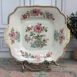 Pode incluir: Um prato quadrado de cer&acirc;mica esbranqui&ccedil;ada com borda ondulada e acabamento dourado. O prato apresenta um desenho floral com flores rosa, vermelhas e azuis e folhas verdes. O prato &eacute; exibido em um suporte de metal decorativo.
