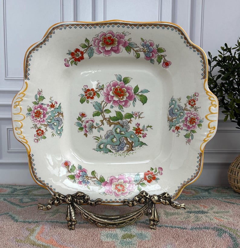 Pode incluir: Um prato quadrado de cer&acirc;mica esbranqui&ccedil;ada com borda ondulada e acabamento dourado. O prato apresenta um desenho floral com flores rosa, vermelhas e azuis e folhas verdes. O prato &eacute; exibido em um suporte de metal decorativo.