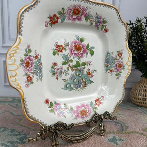 Pode incluir: Prato quadrado de cer&acirc;mica branca com um desenho floral. O prato apresenta flores rosa, vermelhas e azuis com folhas verdes. Possui uma borda dourada e &eacute; exibido em um suporte de metal decorativo.