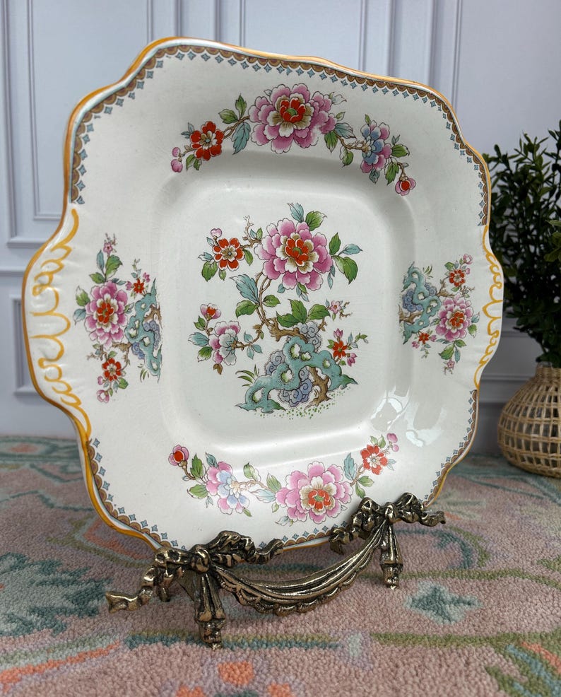 Pode incluir: Prato quadrado de cer&acirc;mica branca com um desenho floral. O prato apresenta flores rosa, vermelhas e azuis com folhas verdes. Possui uma borda dourada e &eacute; exibido em um suporte de metal decorativo.