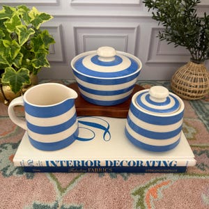 May include: A cream-colored creamer, sugar bowl with lid, and a serving bowl with lid. All pieces have blue and white horizontal stripes. The items are arranged on a decorative book with the title "Interior Decorating".