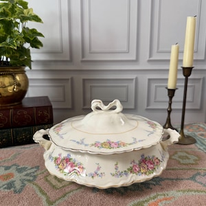 May include: Cream-colored, lidded serving dish with floral patterns in pink, yellow, and blue. The dish has a gold trim and a bow-shaped handle on the lid. Two small handles are on the sides. A vintage piece.
