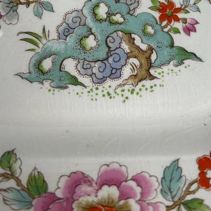 Pode incluir: Prato de cer&acirc;mica branca com um desenho floral. O prato apresenta uma grande pe&oacute;nia rosa com folhas verdes e azuis, e um desenho turquesa em forma de nuvem com ramos castanhos. O prato tem um aspeto rachado e envelhecido.
