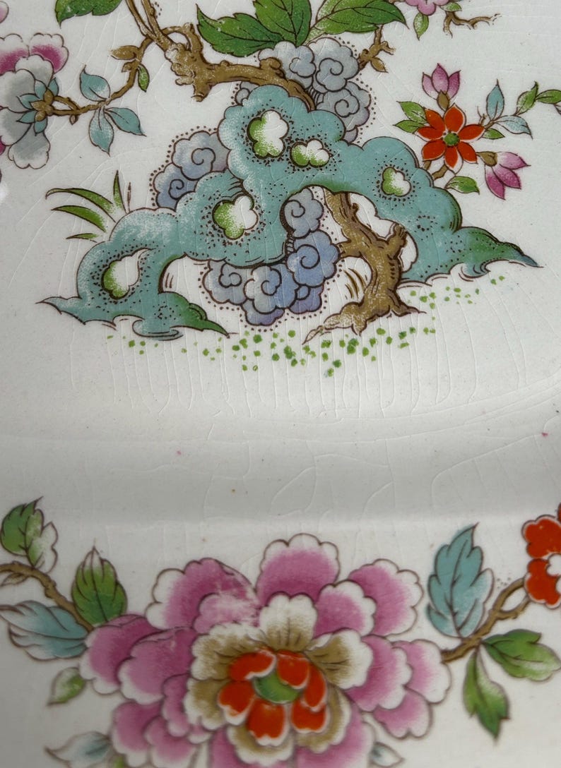 Pode incluir: Prato de cer&acirc;mica branca com um desenho floral. O prato apresenta uma grande pe&oacute;nia rosa com folhas verdes e azuis, e um desenho turquesa em forma de nuvem com ramos castanhos. O prato tem um aspeto rachado e envelhecido.