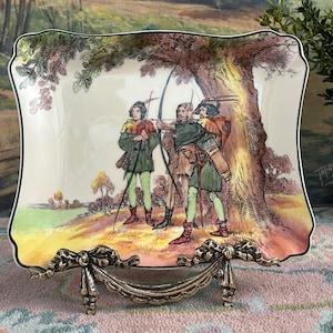 May include: A rectangular decorative plate with a detailed illustration of archers under a tree. The plate has a scalloped edge with a black trim and is supported by a decorative metal stand. The illustration features figures in green and brown clothing.
