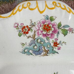 Pode incluir: Um prato de cer&acirc;mica branco com borda ondulada e design floral. O design apresenta flores rosa e vermelhas com folhas verdes e um elemento decorativo azul e verde. A borda &eacute; adornada com uma linha ondulada amarela.