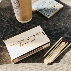 May include: A wooden matchbox with the inscription "You light up my life Frank xxx". The matchbox is on a wooden surface with a bundle of matches next to it.