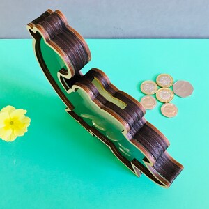 May include: A wooden piggy bank shaped like a cartoon dinosaur. The dinosaur is green and has a brown wooden frame. The piggy bank is sitting on a green surface with coins scattered around it.