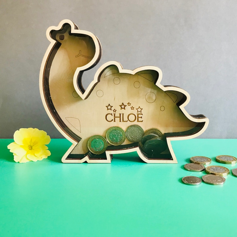 May include: Wooden dinosaur piggy bank with a slot on the back for coins. The dinosaur is light brown and has a name, "CHLOE", engraved on the side. There are coins inside the piggy bank.