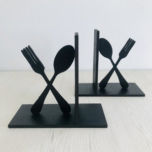 Puede incluir: Un par de sujetalibros de madera negra con forma de tenedor y cuchara. El tenedor y la cuchara est&aacute;n cruzados.