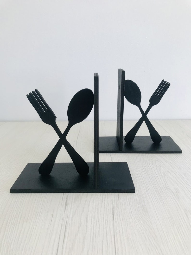 Puede incluir: Un par de sujetalibros de madera negra con forma de tenedor y cuchara. El tenedor y la cuchara est&aacute;n cruzados.