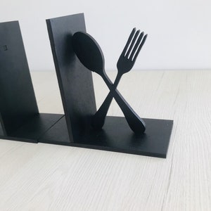 Puede incluir: Dos sujetalibros de madera negra con forma de tenedor y cuchara, cruzados entre s&iacute;.