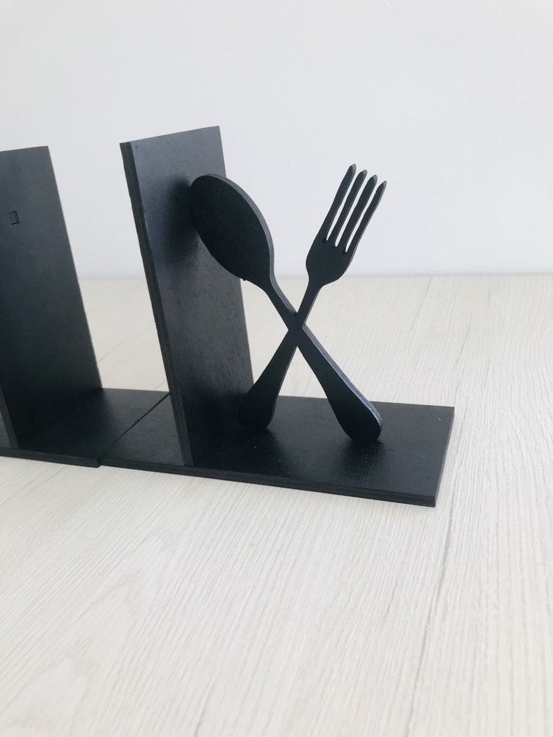 Puede incluir: Dos sujetalibros de madera negra con forma de tenedor y cuchara, cruzados entre s&iacute;.