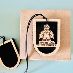 May include: A shield-shaped wooden hanging ornament with a black centre and a light wood border. The ornament features an engraved graphic of a superhero figure and the text "SUPER DAD SUPER DRIVER" in a black font. A black cord is attached to the top.