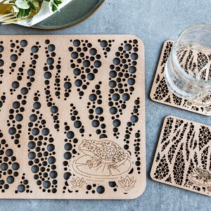 May include: Set of three wooden coasters featuring a frog design. The coasters have a pattern of circles and lines, and each coaster depicts a frog sitting on a lily pad. The coasters are made from light brown wood.