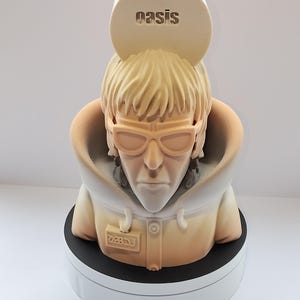 May include: A light tan sculpture of a person wearing sunglasses and a hooded jacket. The sculpture has a white hood and a black base. The word "oasis" is embossed on a circular piece above the head.