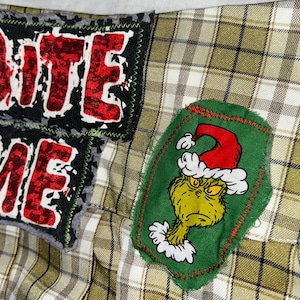 Puede incluir: Una tela a cuadros con tres parches. Dos parches muestran "BITE ME" en letras rojas sobre un fondo negro. El tercer parche es verde con un dibujo del Grinch con un gorro de Papá Noel. También se ve un copo de nieve.