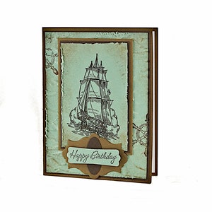May include: A handmade birthday card with a vintage nautical theme. The card features a drawing of a sailing ship on a blue background with a brown border. The text "Happy Birthday" is written on a brown label.