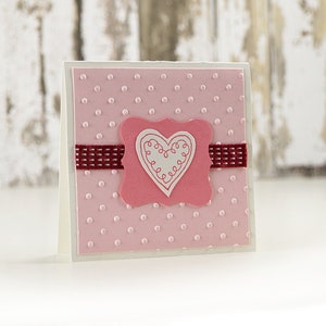 May include: A pink card with white polka dots and a pink heart with a white outline in the center. The heart is surrounded by a white frame with a burgundy ribbon.
