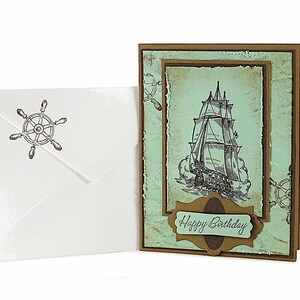 May include: A handmade birthday card with a nautical theme. The card features a vintage illustration of a sailing ship on a blue and green background. The card is decorated with a brown paper border and a brown paper tag with the text "Happy Birthday".