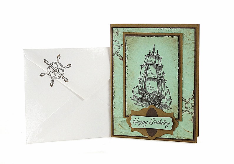 May include: A handmade birthday card with a nautical theme. The card features a vintage illustration of a sailing ship on a blue and green background. The card is decorated with a brown paper border and a brown paper tag with the text "Happy Birthday".