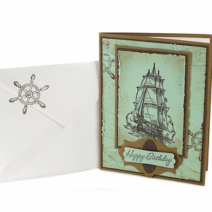 May include: A handmade birthday card with a vintage nautical theme. The card features a drawing of a ship with sails, a brown banner with the text "Happy Birthday", and a blue and green background.