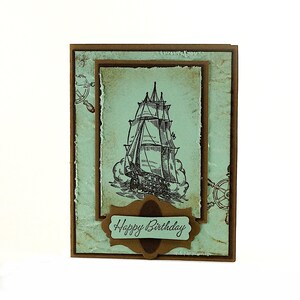 May include: A brown card with a blue background and a drawing of a ship. The card has a brown label with the text "Happy Birthday" on it.