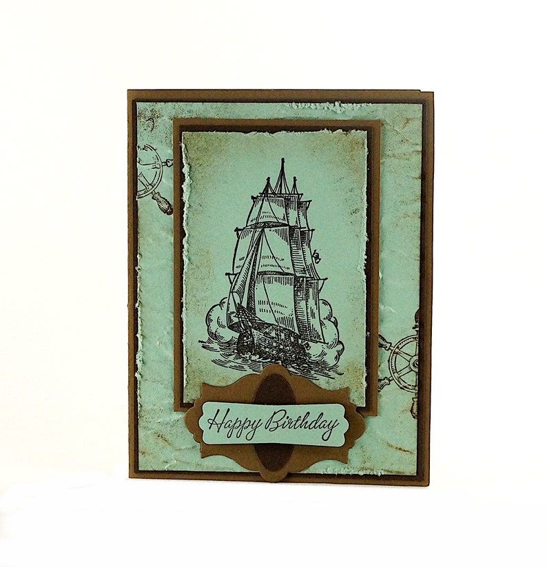 May include: A brown card with a blue background and a drawing of a ship. The card has a brown label with the text "Happy Birthday" on it.