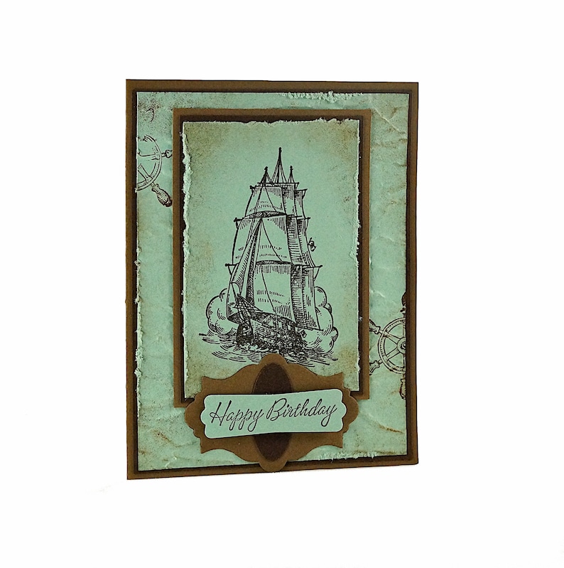 May include: A light blue card with a brown border and a vintage-style illustration of a ship with sails. The card has a brown banner with the text "Happy Birthday" on it.