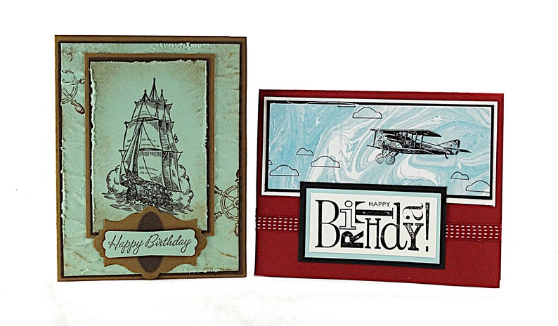 May include: Two handmade birthday cards. The first card features a vintage ship illustration with a blue and brown color scheme and the text "Happy Birthday". The second card features a vintage airplane illustration with a red, blue, and black color scheme and the text "Happy Birthday!"
