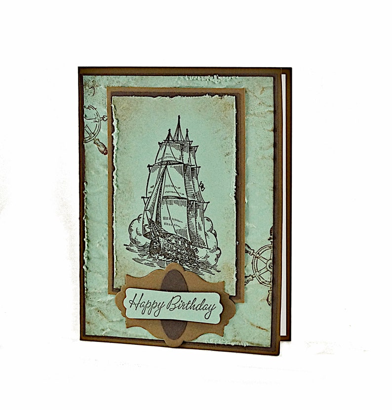 May include: A handmade birthday card with a vintage nautical theme. The card features a drawing of a sailing ship on a blue background with a brown border. The text "Happy Birthday" is written on a brown label.