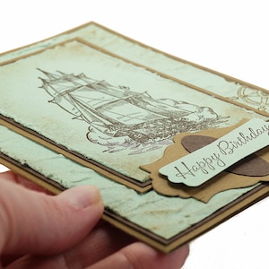 May include: A handmade birthday card with a vintage nautical theme. The card features a brown and white illustration of a sailing ship on a blue and green background. The card has a brown label with the text "Happy Birthday" in cursive.