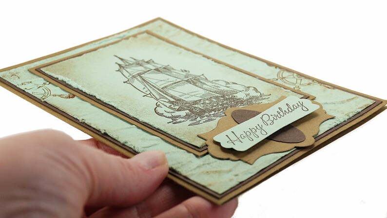 May include: A handmade birthday card with a vintage nautical theme. The card features a brown and white illustration of a sailing ship on a blue and green background. The card has a brown label with the text "Happy Birthday" in cursive.