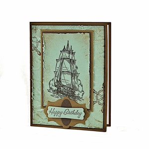 May include: A handmade birthday card with a vintage nautical theme. The card features a drawing of a sailing ship on a blue background with a brown border. The text "Happy Birthday" is written on a brown label.