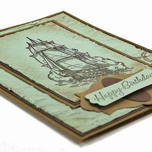 May include: A handmade card with a vintage nautical theme. The card features a brown ship illustration on a light blue background with a brown border. The card has a brown label with the text "Happy Birthday" on it.