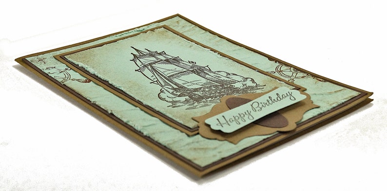 May include: A handmade card with a vintage nautical theme. The card features a brown ship illustration on a light blue background with a brown border. The card has a brown label with the text "Happy Birthday" on it.