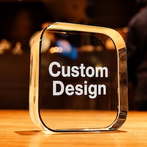 Op de afbeelding: Een heldere glazen prijs met een rechthoekige vorm, afgeronde hoeken en een goudkleurige rand. De woorden "Custom Design" zijn in het wit gedrukt. De prijs staat op een houten oppervlak.