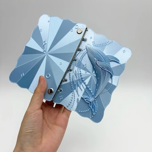 May include: A light blue notebook with a scalloped edge and a three-dimensional whale relief on the cover. The cover features a geometric sunburst pattern with small star accents and a school of fish, held together by two silver metal binder rings.