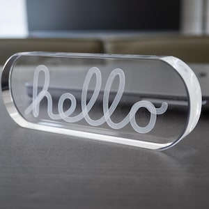 Könnte beinhalten: Ein klarer, rechteckiger Acrylblock mit abgerundeten Kanten. Das Wort "hello" ist in einer Schreibschrift auf die Vorderseite geätzt. Der Block liegt auf einer dunklen Oberfläche mit verschwommenem Hintergrund.