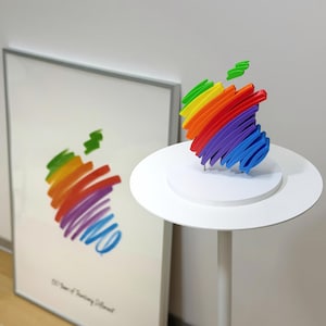 Könnte beinhalten: Eine dreidimensionale Skulptur eines Apfel-Logos in Regenbogenfarben mit geschichteten, pinselstrichartigen Segmenten. Die Skulptur steht auf einem runden weißen Sockel. Im Hintergrund ist ein gerahmter Druck desselben Apfel-Logos zu sehen.