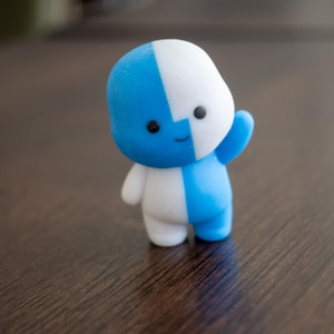 Puede incluir: Una pequeña figura estilizada con un diseño bicolor en azul brillante y blanco. El personaje tiene una cabeza redondeada, dos pequeños ojos negros, una línea curva sencilla para la boca y un brazo levantado en un gesto de saludo.
